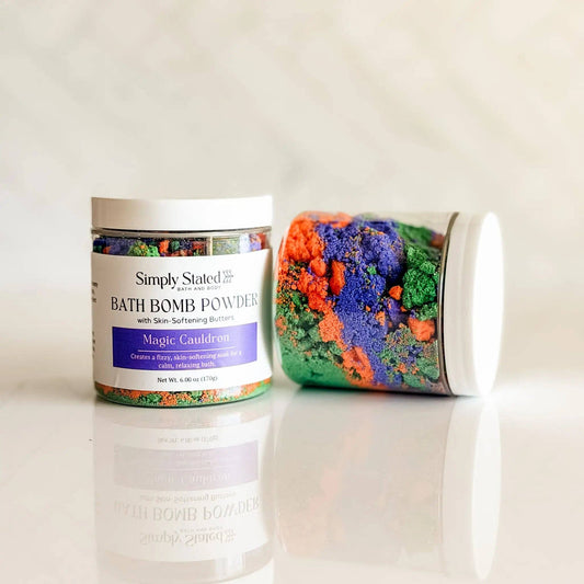 Two white jars of Simply Stated Magic Cauldron bath bomb powders, one standing upright and the other lying on its side, showcasing colorful crystals inside, set against a light marble background.