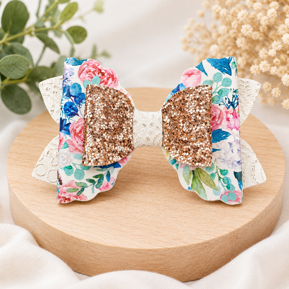A floral hair bow with glittery rose centers, displayed on a round wooden stand with soft fabric and dried flowers in the background.