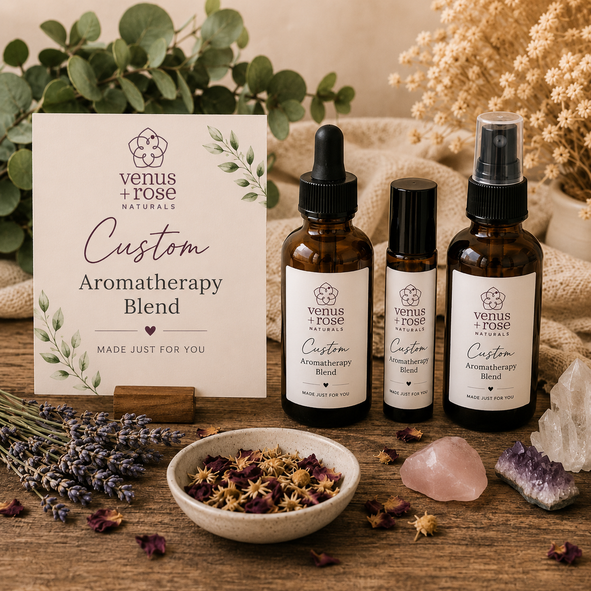 Three amber glass aromatherapy bottles and a matching branded card from Venus + Rose Naturals arranged on a wooden surface with dried flowers, crystals, and greenery in a styled product scene.