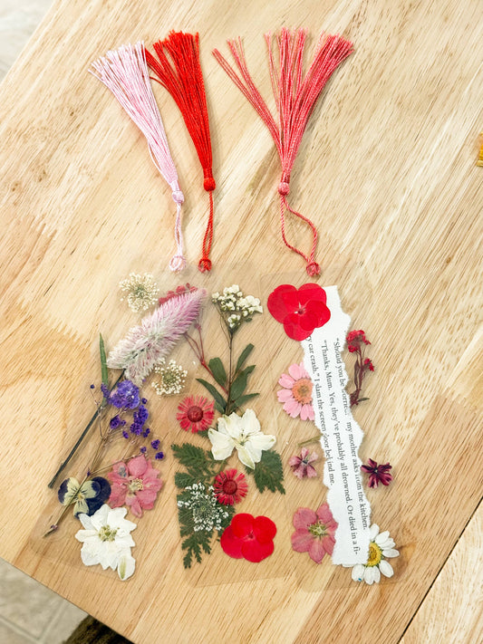 A clear acrylic keepsake tag decorated with pressed flowers, green leaves, a white paper strip with printed text, and two tassels at the top, one red and one pink, arranged on a light wood surface.