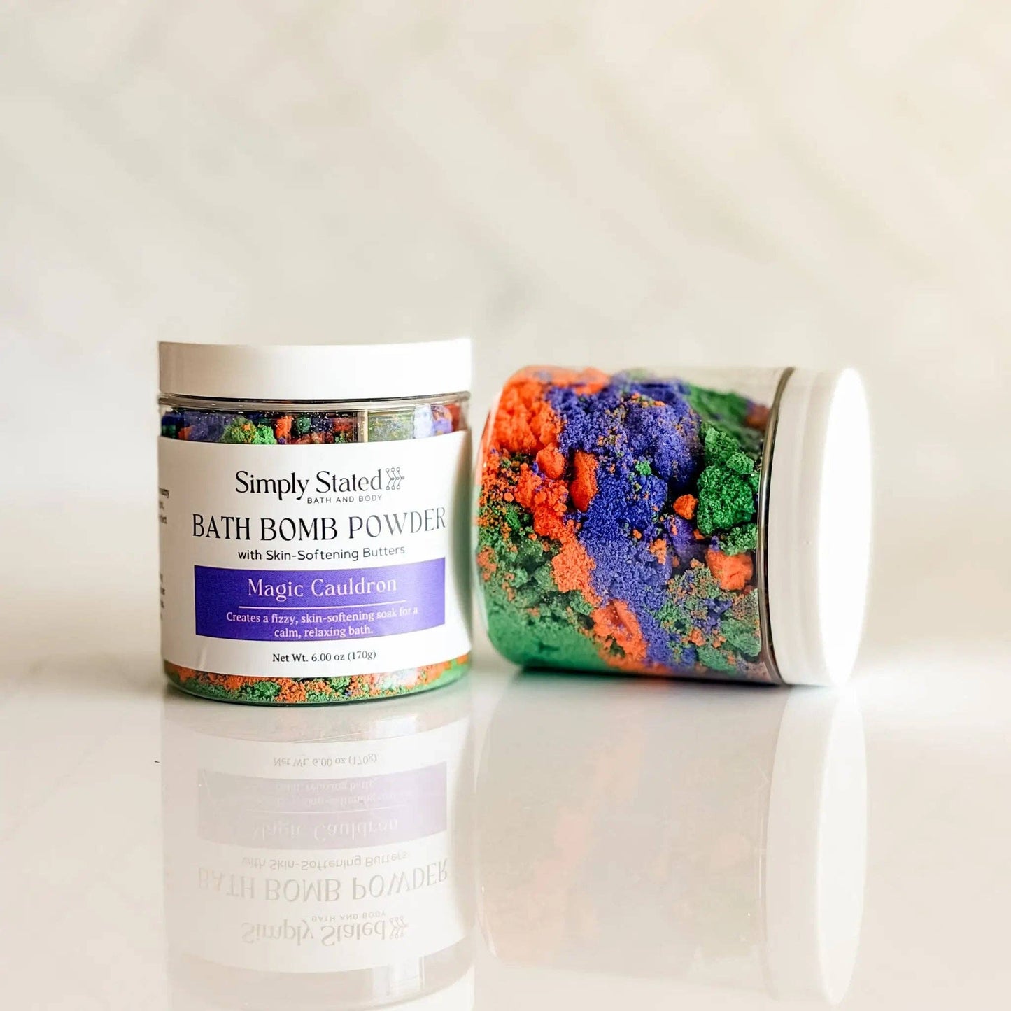 Two white jars of Simply Stated Magic Cauldron bath bomb powders, one standing upright and the other lying on its side, showcasing colorful crystals inside, set against a light marble background.