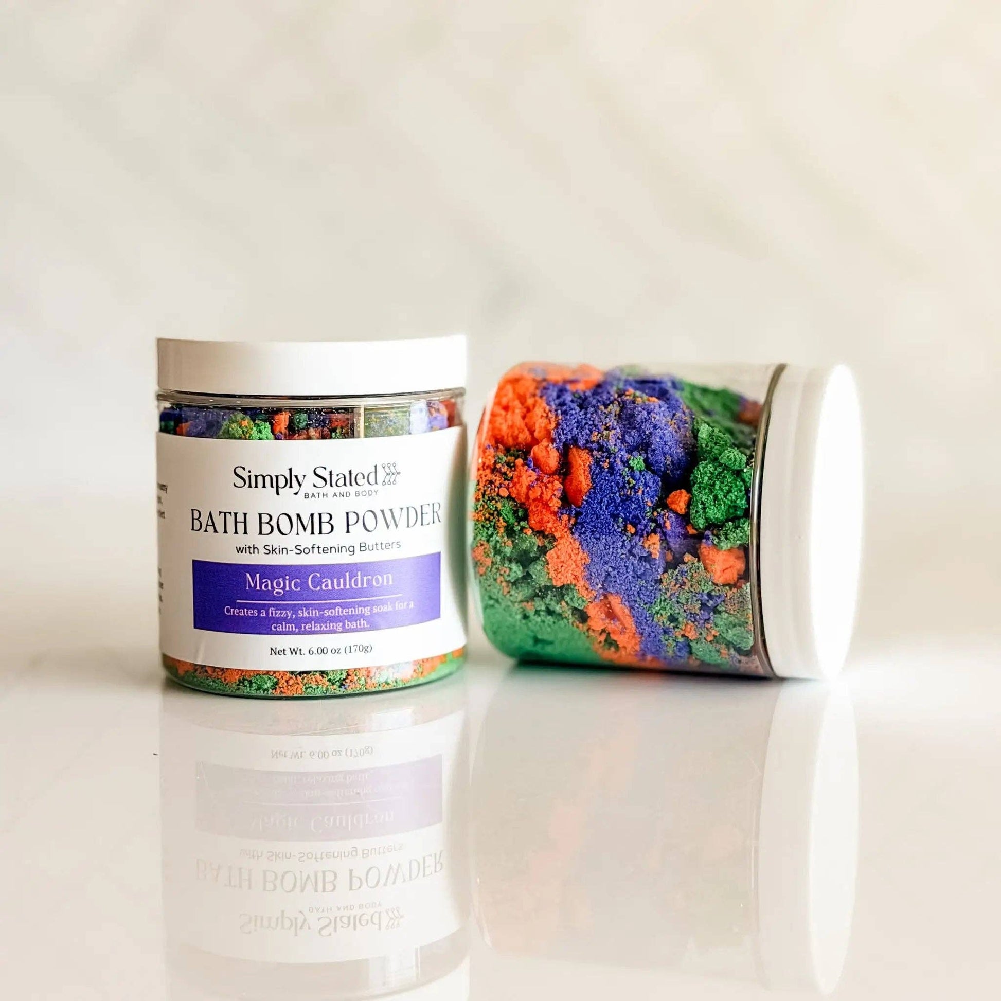 Two white jars of Simply Stated Magic Cauldron bath bomb powders, one standing upright and the other lying on its side, showcasing colorful crystals inside, set against a light marble background.