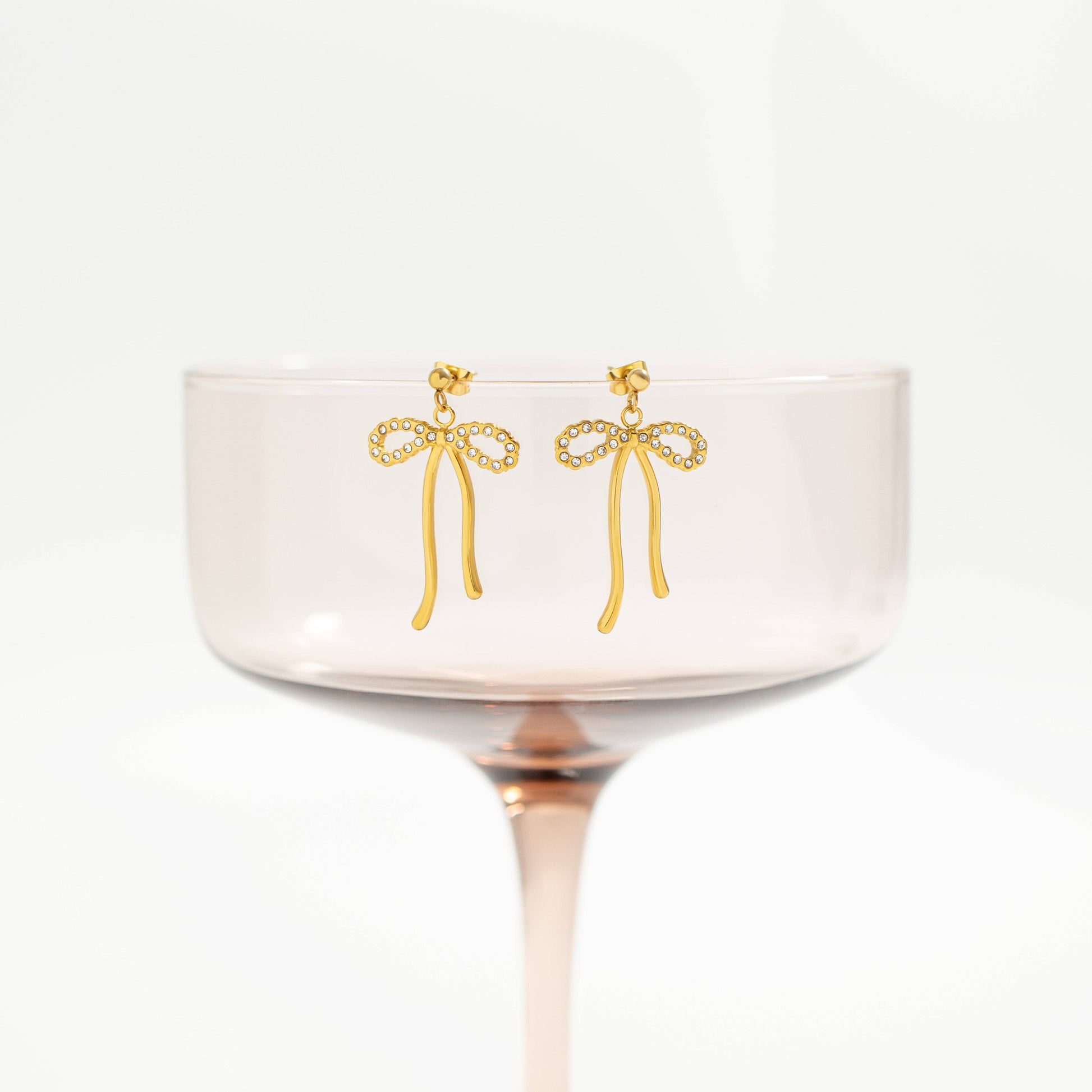 A pair of gold bow-shaped dangle earrings with small clear stones along the bow edges, displayed on the rim of a pale pink glass coupe against a white background.