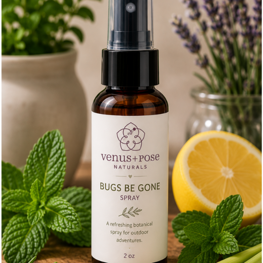 Venus + Pose Naturals 'Bugs Be Gone' spray bottle displayed on a rustic wood slice, surrounded by fresh botanical ingredients including mint leaves, rosemary, lemongrass, a lemon half, and potted plants in the background.