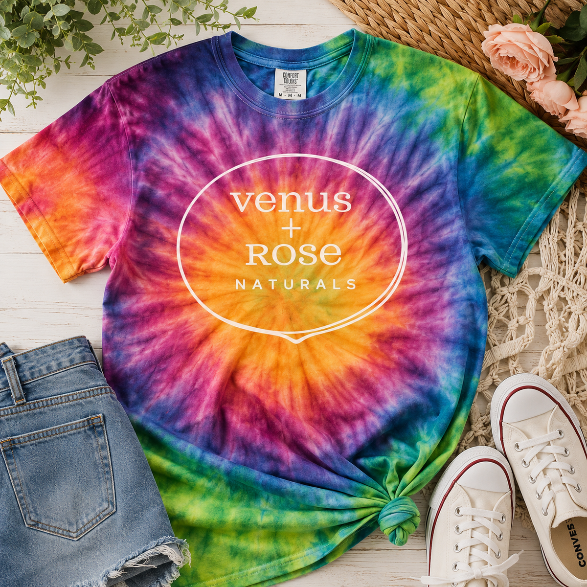 A colorful tie-dye t-shirt with a spiral pattern in purple, blue, green, orange, and pink, featuring a white circular logo that reads 'venus + rose NATURALS'. The shirt is styled in a flat-lay arrangement with blue denim shorts, white canvas sneakers, and a woven straw hat on a white wood background.