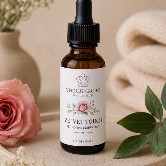 A 1 fl oz amber glass dropper bottle of Venus+Rose Naturals Velvet Touch Personal Lubricant, styled on a beige surface with a pink rose, green leaves, and a beige towel in the background.