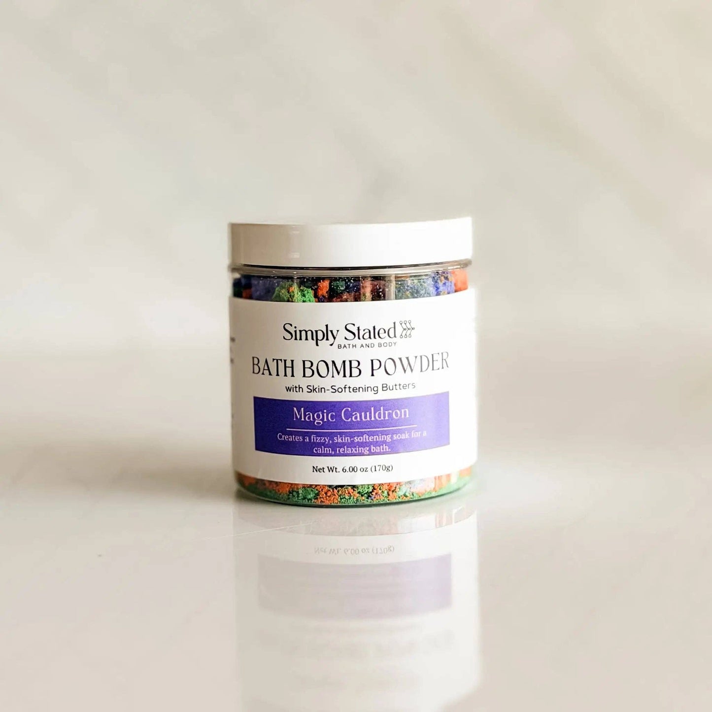 A jar of Simply Stated 'Magic Cauldon' bath bomb powder, featuring a white label with a purple accent band and a white lid, set against a clean, light gray reflective background.