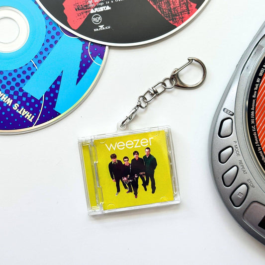 A Weezer acrylic keychain charm in the yellow album art style, attached to a silver metal clasp, displayed on a white surface with CD-ROMs and a cassette tape partially visible as props.