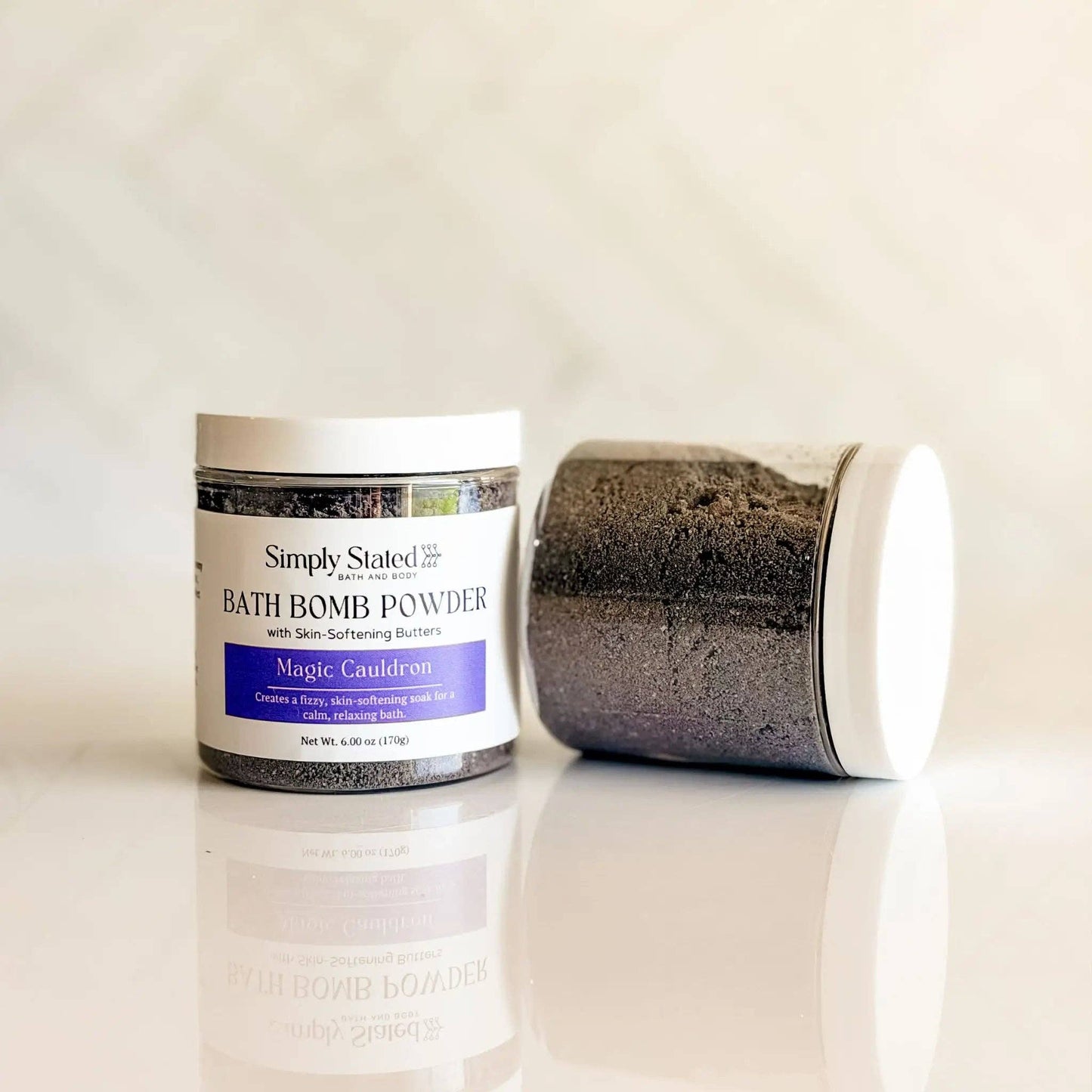 A jar of Simply Stated Apothecary Magic Cauldron bath bomb powder stands upright while another jar lies on its side, both showing the dark purple powder inside white jars with white lids, set against a light marble background.