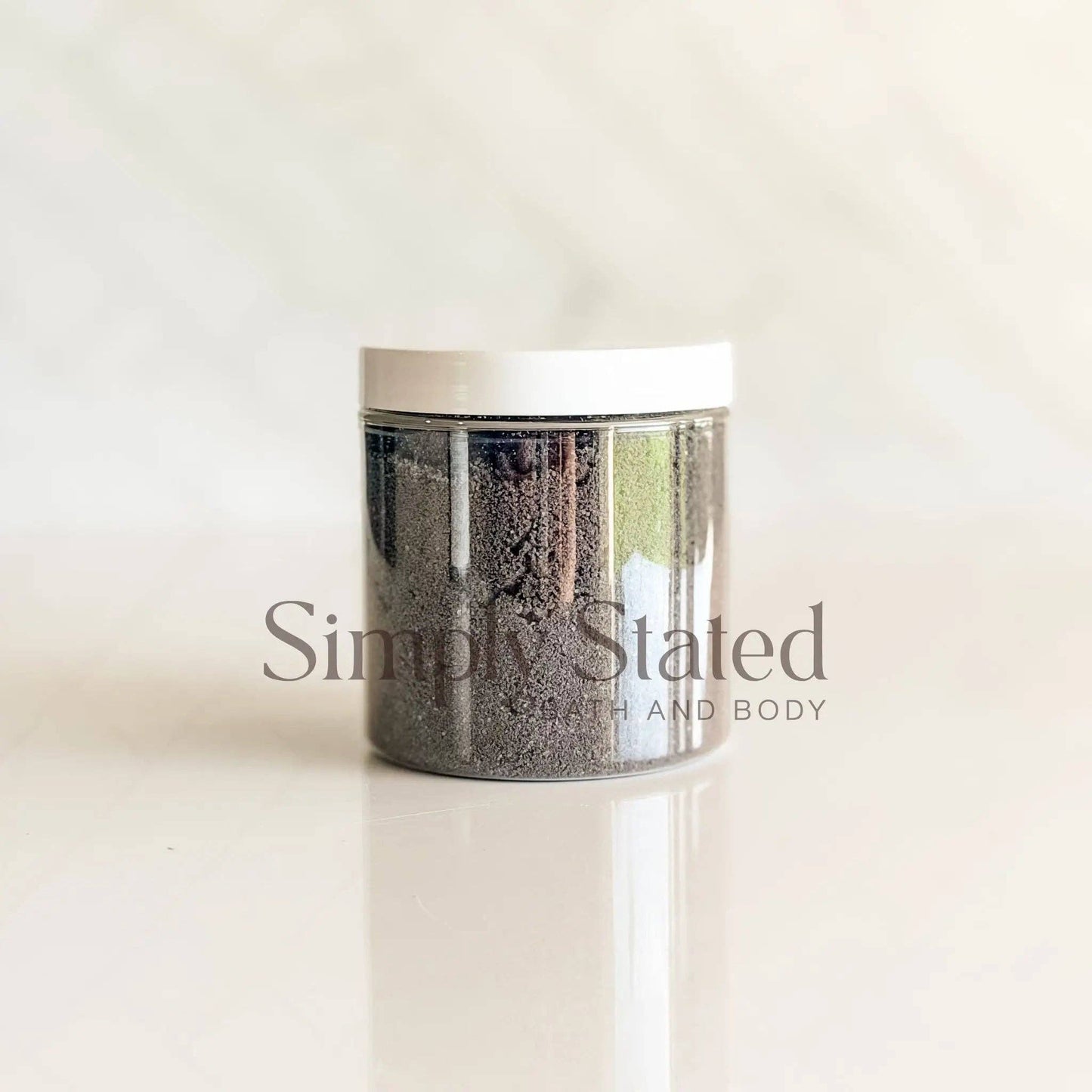 A clear cylindrical jar filled with dark gray exfoliating scrub, featuring a white lid, set against a light beige marble background with a reflection below. The brand name 'Simply Stated' is watermarked across the center.