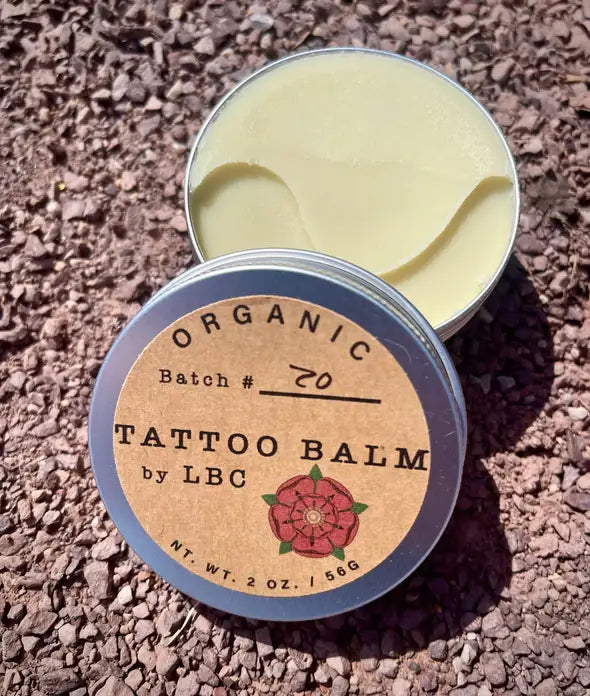 An open round tin of organic tattoo balm showing the pale yellow balm inside, with the lid placed in front. The lid label reads 'ORGANIC TATTOO BALM by LBC' with a small rose illustration, photographed outdoors on gravel.