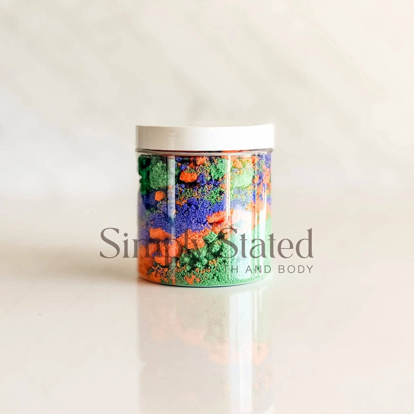 A clear plastic jar filled with colorful textured bath salts in shades of green, orange, and blue, topped with a white lid, set against a light beige marble background with a faint reflection.