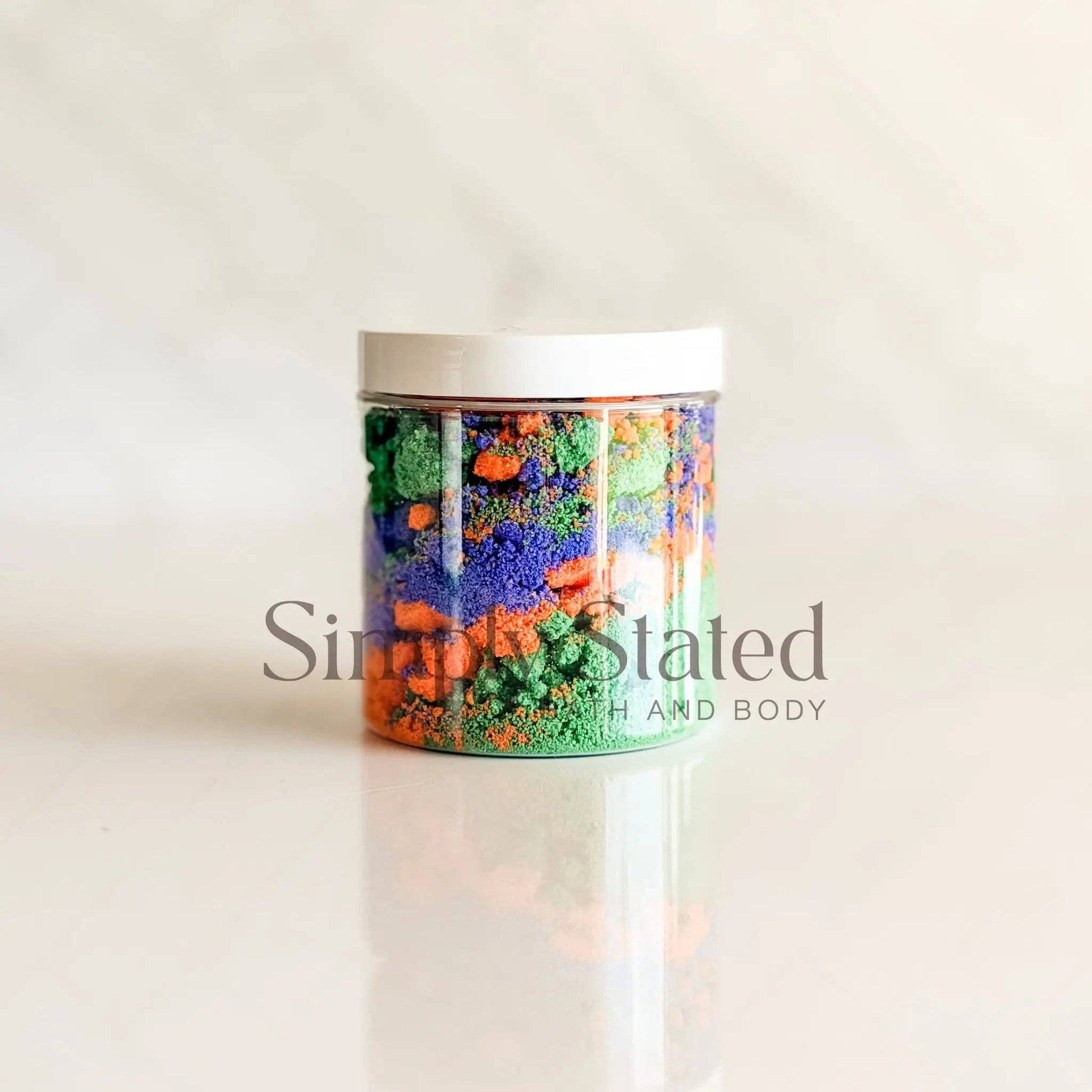 A clear plastic jar filled with colorful textured bath salts in shades of green, orange, and blue, topped with a white lid, set against a light beige marble background with a faint reflection.