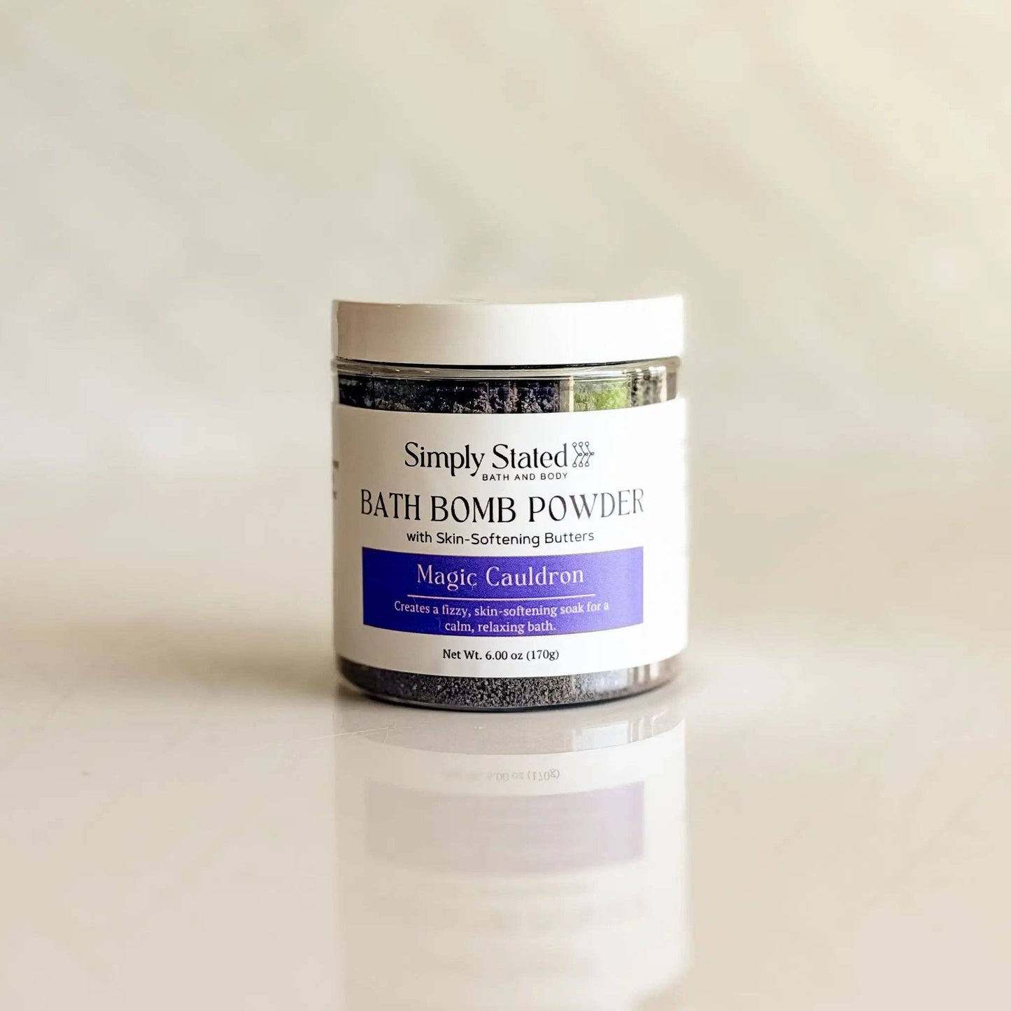 A jar of Simply Stated Bath and Body 'Magic Cauldron' bath bomb powder, featuring a white label with a purple accent band, set against a soft, light beige background with a subtle reflection.